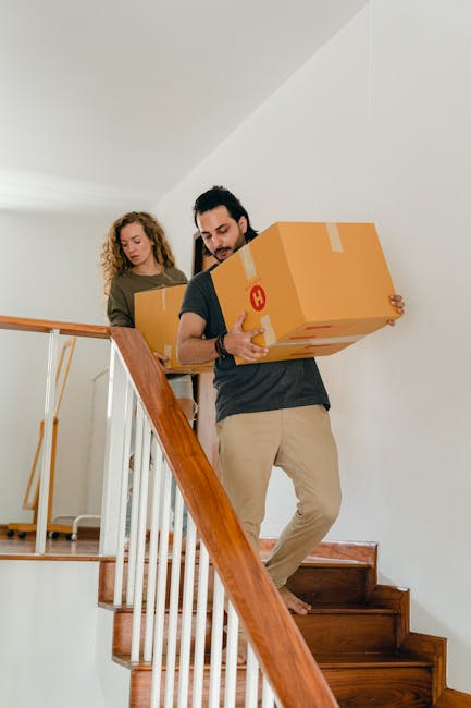 A woman with long dark hair, dressed in a beige top and jeans, is carrying a large cardboard box up a narrow indoor staircase during a home relocation. The staircase is carpeted in dark fabric, with a
