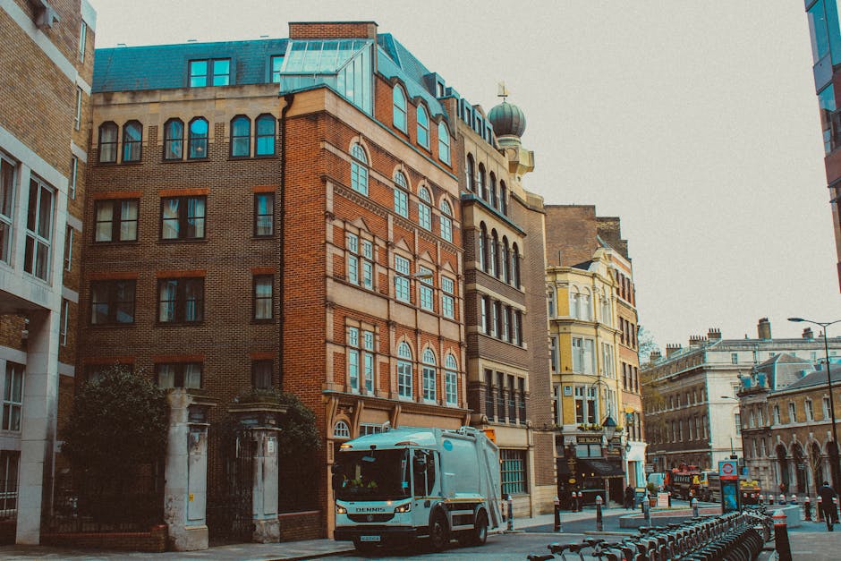 The image shows a city street scene with a white moving van parked in front of a row of multi-storey buildings featuring a mix of brick and stucco facades, some with large arched windows and ornate ar