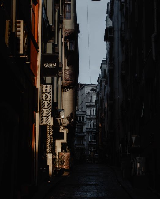 A narrow cobblestone street in an urban residential area during daytime, lined with multi-storey buildings featuring various architectural styles. On the left side, there are several commercial signs